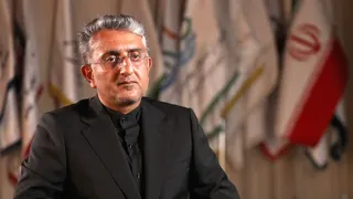 A man wearing a black shirt and jacket looks at the camera. He is sitting down with flags behind him, one of which is the Iranian flag.