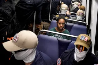 A Black woman sits surrounded by neo-fascists in white masks.