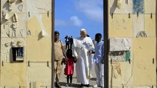 Pope Leo holds hands with a girl wearing a red dress and a man in blue shirt and white trousers. A women in a beige outfit stands alongside them at a doorway. Behind them are blue skies and men dressed in black