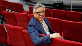 Jin Mingri is wearing a blue blazer and white collared shirt, sitting in a row of red upholstered seats in a church auditorium