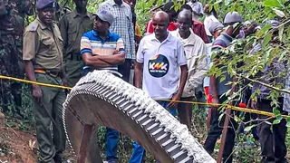 A crowd, including police officers behind yellow tape looking on at a giant metal ring which fell from space on to farmland in Mukuku, Kenya