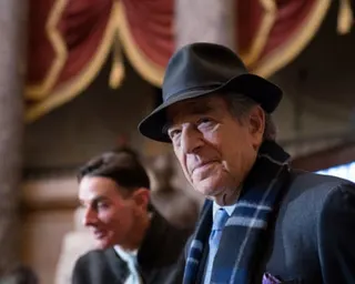 man in a fedora hat, suit and striped scarf stands in an ornate building with blurred figures behind