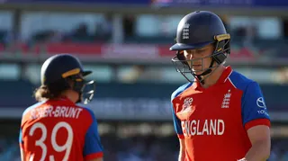 Amy Jones (right), wearing a red England shirt with blue short sleeves and a dark blue England helmet, looks downcast as she leaves the field, with Nat Sciver-Brunt (left) entering the field in the background to replace her