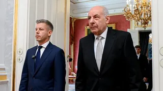 Peter Magyar and Tamas Sulyok walking through a door. A chandelier hags in the room behind them, which also has a room with red-coloured wallpaper.
