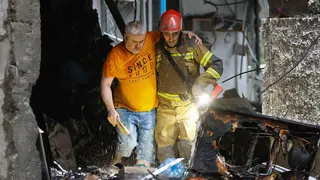 Man in an orange T-shirt helped from a ruined building by an emergency worker