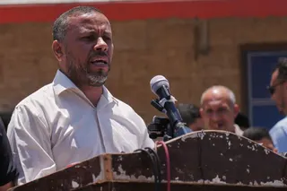 A man in glasses speaks into a cluster of microphones at an outdoor press conference, with people standing behind him
