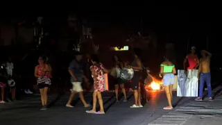 People protest against power outages in Havana on 7 July 2026. One person can be seen holding a large pot while another is banging a metal sheet. In the background burning rubbish can be seen on the street.