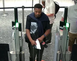 A man in a dark t-shirt walks through airport security barriers while holding documents