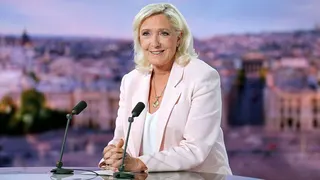 Marine Le Pen, who has shoulder-length blonde hair and is wearing a pastel pink blazer with a white top, smiles as she sits at a sdesk in a French TV studio in front of two microphones, following the verdict in her appeal trial on Tuesday