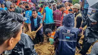 A large group of volunteers and aid workers stand in deep mud during heavy rain, working together at the site of a landslide by a school at a refugee camp in Cox's Bazar