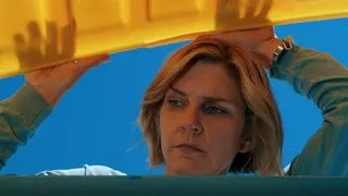 Rhea Seehorn seen from below opening a large yellow lid of a large container and looking inside, in a still from Pluribus