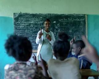 A female teacher stands in front of a blackboard teaching a class of children.