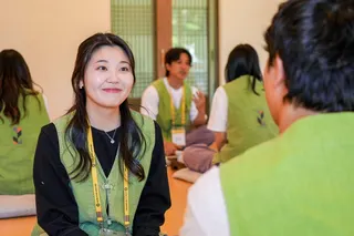 A participant wearing a light green vest and yellow lanyard sits on a floor cushion in a temple hall, facing another participant. Several others are visible in the background