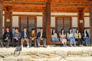 A group of five men and five women wearing lanyards sit along a wooden porch of a traditional Korean temple with large timber beams and lattice doors. The group is spread across the veranda in bright daylight, with a stone foundation in the foreground and a bag resting near the front edge.