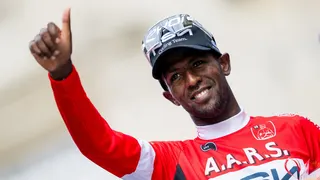 Biniam Girmay, wearing a red long-sleeved cycling jersey and clear sunglasses on top of a blue baseball cap, smiles as gives a thumbs up gesture with his right arm raised in front of him