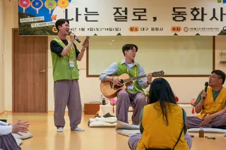 Participants sit and perform music at the temple dating retreat - one person stands holding a microphone, while another sits playing an acoustic guitar. Other attendees sit on floor cushions in a circle. A large banner with Korean text hangs behind the group.