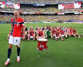 Erling Haaland prepares to bang a drum as his teammates sit on the ground in rowing formation