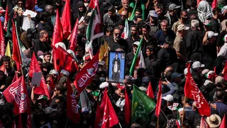 Mourners gather for the burial of Iranian Supreme Leader Ali Khamenei in Mashhad. Many are carrying red flags with Arabic script and in the centre a man holds a picture of a sitting Khamenei, with his son Mojtaba standing above him.