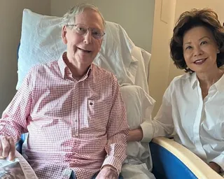 a man sits in a hospital bed with his wife holding a newspaper