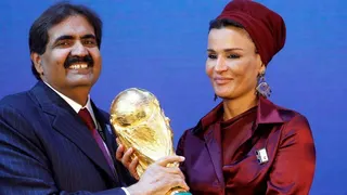 Sheikh Hamad grins as he grips the trophy with his wife. He is dressed in a blue pin strip suit and his wife is in a maroon coloured outfit.