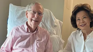 An elderly man in a pink checkered shirt sits in a hospital bed, smiling, with a newspaper in his lap. A smiling woman in a white blouse sits beside him in a hospital room.
