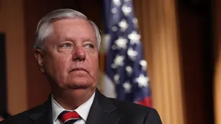 Lindsey Graham, a man with short, grey hair, wears a black suit with a white shirt and a red and black tie. He is looking to the right. The background behind him is blurred, with a hanging American flag.