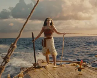 Catherine Laga'aia as Moana in traditional Polynesian clothing stands on a wooden raft holding a rope and paddle at sea