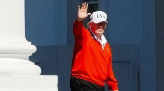 President Donald Trump, wearing a white USA cap and a red golfing sweater, waves as he passes by white pillars outside the White House