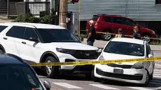 Two white cars that collided are behind yellow police tape. The windscreen of one car has bullet holes in it