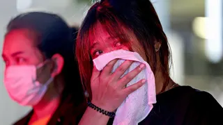 Two women stand, their faces lit by emergency services lights, one wearing a face mask and the other holding a white cloth over her mouth and nose.
