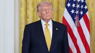 Donald Trump, in a dark navy suit and yellow tie, stands with his hands at his sides and a neutral expression. Behind him are gold curtains in the White House and an American flag.