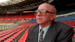 Nobby Stiles sits in the stands at Wembley. He is wearing a suit and glasses with a yellow tint.