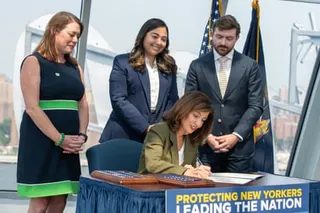 two women and a man stand behind a woman signing a document while seated at a desk