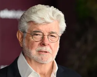 George Lucas with white hair and a grey beard wearing glasses and a dark jacket