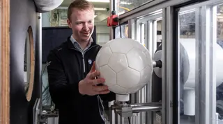 A man wearing a black zip-up jumper holds a white football on a contraption in a lab. He has blonde hair and is smiling slightly.