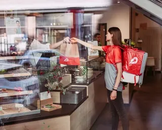 A Delivery Hero worker wearing a red backpack receives a takeaway bag from a restaurant worker at a counter