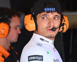 Lando Norris wearing orange headphones and a blue sensor headband looks upward in a garage.