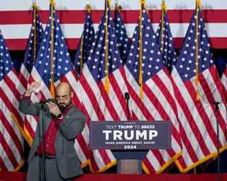 A man gestures at a podium with Trump 2024 campaign signage and multiple American flags in the background