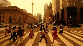 People walk on a crossing in a road downtown Toronto. There are buildings in the background and sky is orange from smoke.