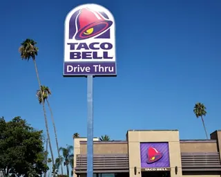 Pink, purple and yellow Taco Bell logo on a drive-thru sign, and a restaurant, amid very high palm trees.