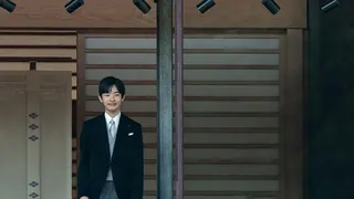 Prince Hisahito smiles and stands behind a window. He is wearing a black suit.