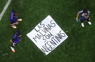 A banner saying "Las Malvinas Son Argentinas" is on the ground of a pitch with three players standing around it