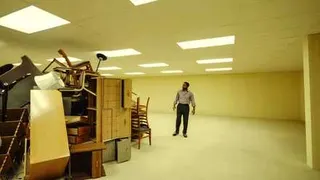 A Black man (Chiwetel Ejiofor) in a button-down shirt and black slacks stands in a huge room with the walls, floor, and ceiling all painted the same sallow yellow. He's examining a huge pile of jumbled furniture, the only thing in the room.