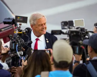 Smiling man in suit with red tie surrounded by reporters.