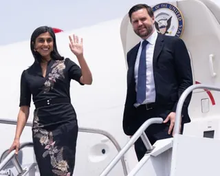 woman in floral dress waves and man in suit smiles as they step outside plane