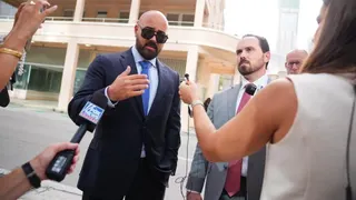 Joseph McBride, bald and with a beard and mustache and wearing sunglasses and a blue suit, shirt and tie, and Thomas Maniotis, with short cropped brown haior and a beard and mustache, with a grey suit and red tie, speak into microphones surrounded by media