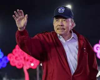 Daniel Ortega in a red jacket and navy cap waves his hand at an evening event with colorful lights behind him