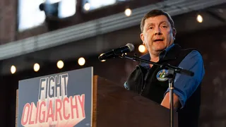A man wearing a blue collared shirt and a black vest, Troy Jackson, speaks into a microphone from behind a podium that features a sign reading "FIGHT OLIGARCHY."
