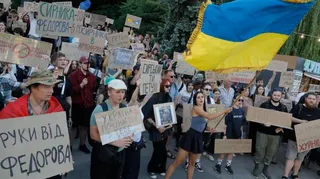 Dozens of people, mostly young, holding cardboard signs in a protest, as one woman waves a large Ukrainian flag