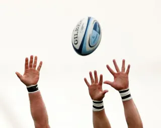 Three hands reach up toward a rugby ball suspended in the air against a pale sky